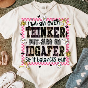 May include: Cream-colored t-shirt with the text "I'm an over thinker but also an IDGAF-er so it balances out." The design includes pink checkered borders, flowers, butterflies, and yellow bows. The shirt has a crew neck.