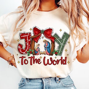 May include: A white t-shirt with a red, green, and gold "JOY" graphic with a red bow and a nativity scene in the center. The text "To The World" is below the graphic.