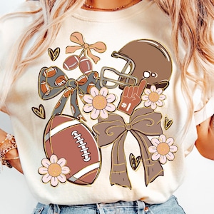 Op de afbeelding: Crèmekleurig T-shirt met een voetbalthema-afbeelding. Het ontwerp bevat een voetbal, helm en een #1 vinger, samen met bloemen- en strikaccenten. Het shirt is een casual stijl.