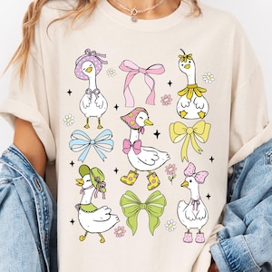 Puede incluir: Una camiseta de color crema con un gr&aacute;fico de seis gansos de dibujos animados que llevan sombreros y lazos. Los gansos son blancos con picos y patas amarillas. Los sombreros son de varios colores y estilos, incluyendo un sombrero de flor rosa, un sombrero verde con un lazo y un sombrero azul con un lazo. Los lazos tambi&eacute;n son de varios colores y estilos, incluyendo un lazo rosa, un lazo azul y un lazo amarillo. Los gansos est&aacute;n rodeados de peque&ntilde;as flores y estrellas.
