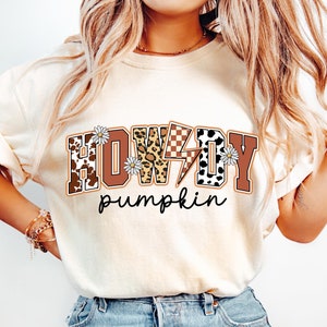 May include: Cream-colored t-shirt with the words "HOWDY pumpkin" in a western-themed design. The word "HOWDY" features cow print, leopard print, and a checkered pattern. Daisies and a lightning bolt accent the design.