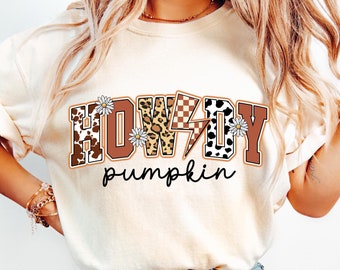 Peut inclure: T-shirt crème avec l'inscription "HOWDY pumpkin" dans un design sur le thème du western. Le mot "HOWDY" présente un imprimé vache, un imprimé léopard et un motif à carreaux. Des marguerites et un éclair ornent le design.