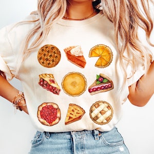 May include: A white t-shirt with a graphic design of various pie slices. The slices are in different colors and patterns, including cherry, pumpkin, and apple.