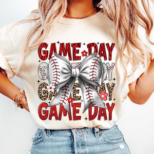 以下が含まれることがあります： 野球をテーマにしたデザインのクリーム色のTシャツ。グラフィックは、野球ボールで作られた大きなリボン、赤い文字で「GAME DAY」、花のアクセントが特徴です。このシャツはブルージーンズと組み合わせています。