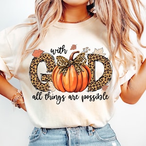 May include: A white t-shirt with a graphic design of the word "GOD" with a pumpkin in the center. The letters are outlined in black and have a leopard print pattern. The pumpkin is orange with a green stem and leaves. The text "with" is above the graphic and the text "all things are possible" is below the graphic.