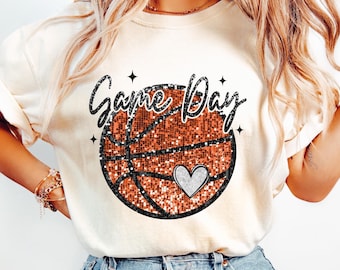 May include: Cream-coloured t-shirt with a basketball design and the words "Game Day" in a glittery font. The basketball is orange and black with a silver heart. The shirt is a casual style.