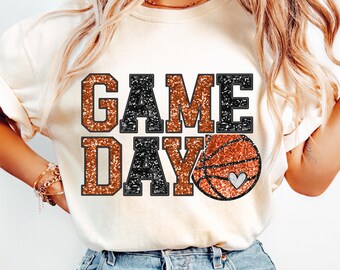 May include: A white t-shirt with the text "GAME DAY" in black and orange glitter with a basketball graphic featuring a small white heart.
