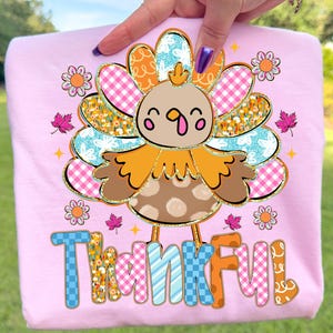 May include: Pink t-shirt featuring a cartoon turkey design with a colorful, patterned tail and the word "THANKFUL" in a playful font. The turkey has a brown body with white polka dots and orange feathers.
