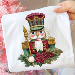 May include: White t-shirt featuring a detailed illustration of a traditional nutcracker soldier. The nutcracker is depicted in shades of red, green, gold, and white, holding a staff and surrounded by festive foliage.