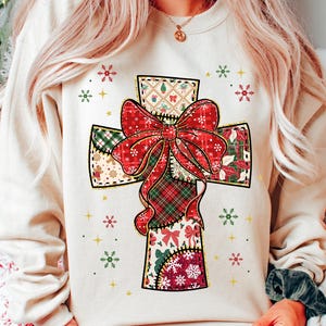Könnte beinhalten: Ein cremefarbenes Sweatshirt mit einem großen Kreuzdesign. Das Kreuz ist mit einer roten Schleife und Patchworkmustern in Grün, Rot und Weiß verziert. Schneeflocken- und Sternakzente umgeben das Kreuz und schaffen ein festliches Urlaubs-Thema.