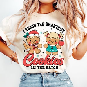 May include: A white t-shirt with the text "I teach the smartest cookies in the batch" in a vintage style font. The text is surrounded by a gingerbread man and a gingerbread girl holding an apple.
