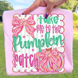 May include: A light pink t-shirt with the phrase "Take me to the Pumpkin Patch" in green and pink lettering. The design includes a bow and a pumpkin illustration, all outlined in gold. The shirt is held up against a green background.