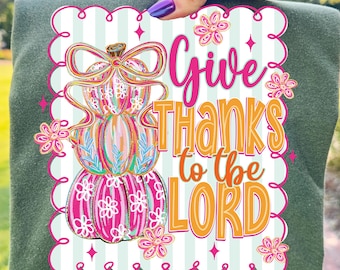 May include: Green long-sleeve shirt with a graphic that says "Give Thanks to the Lord." The graphic features a stack of pumpkins in pink, orange, and blue, with floral accents and a bow.