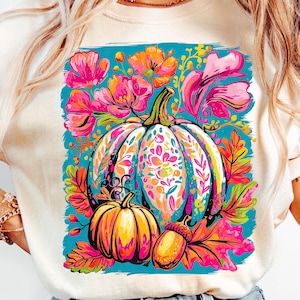 Peut inclure: Sweat-shirt crème avec un motif floral et citrouille vibrant. Le motif comprend une grande citrouille à motifs, une plus petite citrouille et un gland, entourés de fleurs et de feuilles colorées sur fond turquoise.