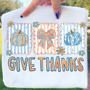 May include: White sweatshirt with a fall-themed design. The design features three framed sections with pumpkins and a bow, set against striped backgrounds. The words "GIVE THANKS" are printed in orange and blue.