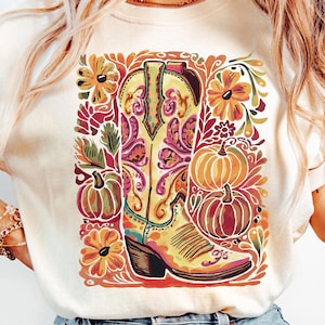 May include: Cream-colored t-shirt featuring a colorful illustration of a cowboy boot, pumpkins, and floral elements. The boot is yellow with pink and purple accents. The design has a fall theme.