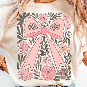 K&ouml;nnte beinhalten: Cremefarbenes Sweatshirt mit einer gro&szlig;en rosa Schleife, umgeben von floralen und Blattillustrationen in Rosa, Grau und Wei&szlig;. Das Design ist auf der Vorderseite zentriert und schafft eine feminine und k&uuml;nstlerische &Auml;sthetik.