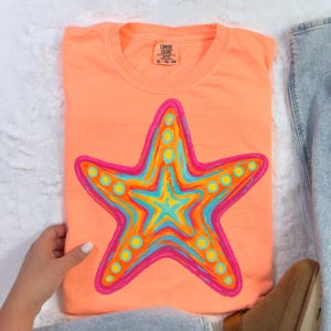 May include: Coral-colored t-shirt featuring a vibrant starfish design. The starfish is outlined in pink and filled with shades of blue, yellow, and orange. The shirt has a crew neck and short sleeves. The tag reads XL-TG-EG.