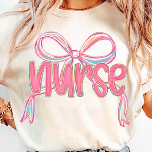 Pode incluir: Camiseta creme com um grande la&ccedil;o rosa e a palavra "Nurse" em rosa. O la&ccedil;o tem listras de arco-&iacute;ris pastel. Uma pe&ccedil;a casual para o dia a dia.