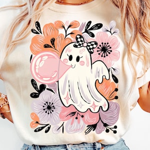 May include: Cream-coloured t-shirt with a cartoon ghost blowing a pink bubblegum bubble. The ghost has a checkered bow and is surrounded by pink, peach, and purple flowers with black leaves. A cute and whimsical design.