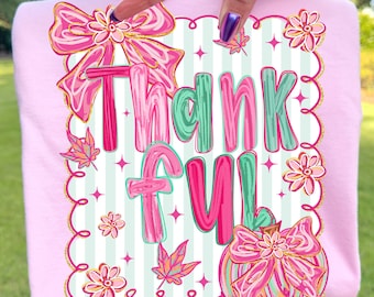 May include: A light pink t-shirt with the word "Thankful" in pink and teal letters. The design includes pink bows, flowers, and leaves with gold accents, set against a striped background.
