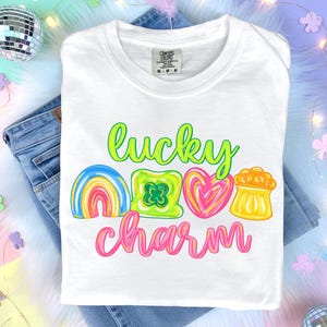 Puede incluir: Camiseta blanca con las palabras "lucky charm" en verde neón y rosa. La camiseta presenta gráficos coloridos de un arcoíris, un trébol, un corazón y una olla de oro. La camiseta está doblada sobre unos vaqueros azules.