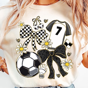 May include: Cream-colored t-shirt featuring a soccer-themed design. The graphic includes a soccer ball, a jersey with the number 7, bows, daisies, and hearts. The design incorporates black, white, and gold accents, creating a playful and sporty aesthetic.