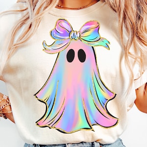 May include: A cream-colored t-shirt featuring a colorful ghost design. The ghost has a rainbow-colored body, black eyes, and a large bow on its head. The design is outlined in gold glitter.
