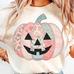 May include: A cream-colored sweatshirt featuring a patchwork pumpkin design. The pumpkin is made of floral and patterned fabrics in pink, green, and cream, with a black face. The design has a stitched appearance, perfect for fall and Halloween.