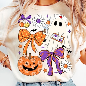 May include: Cream-colored t-shirt featuring Halloween-themed graphics. The design includes a ghost wearing a nurse's cap, a pumpkin, bows, flowers, bats, stars, and a syringe. The overall aesthetic is a blend of spooky and cute.