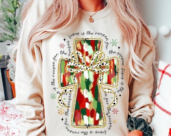 Könnte beinhalten: Ein beiger Pullover mit einem bunten Kreuzdesign und dem Text "Jesus is the reason for the season", der sich um das Kreuz wiederholt.