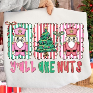 May include: White sweatshirt with a festive Christmas design. It features two pink nutcrackers, a decorated Christmas tree, and the text "Y'all Are Nuts" in pink and green. The design is framed with decorative borders.