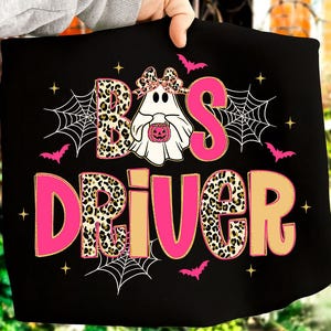 Op de afbeelding: Zwart t-shirt met de tekst "BOO'S DRIVER" in roze en luipaardprint. Het ontwerp bevat een spook met een pompoen, spinnenwebben, vleermuizen en sterren, perfect voor Halloween.