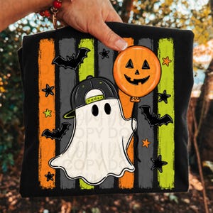 May include: A black t-shirt featuring a graphic of a white ghost wearing a black baseball cap and holding an orange jack-o'-lantern balloon. The background consists of vertical orange, gray, and lime green stripes with black bats and stars.