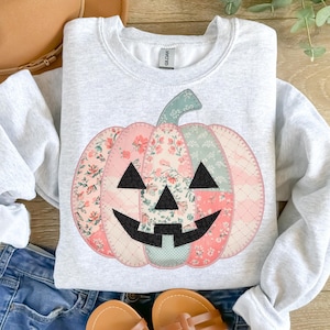 May include: A light gray sweatshirt featuring a patchwork pumpkin design. The pumpkin is made of floral patterned fabric in pink, green, and white, with black triangle eyes and a smiling mouth. The stem is green.