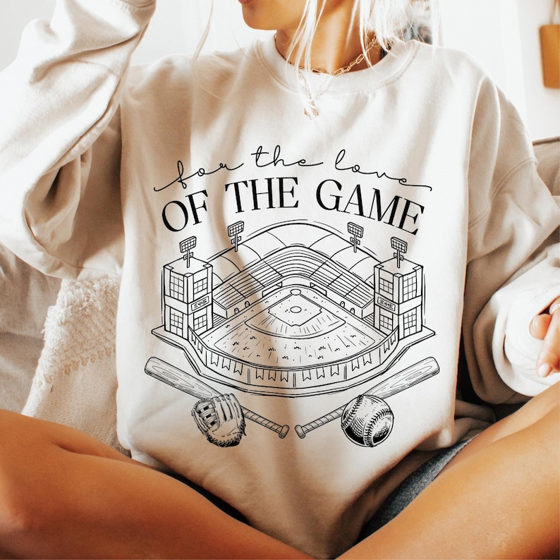 Op de afbeelding: Cr&egrave;mekleurige sweatshirt met een zwart-witte honkbalstadionafbeelding, honkbal, handschoen en knuppels. De tekst "for the love OF THE GAME" staat boven de afbeelding. Een casual, comfortabele stijl.