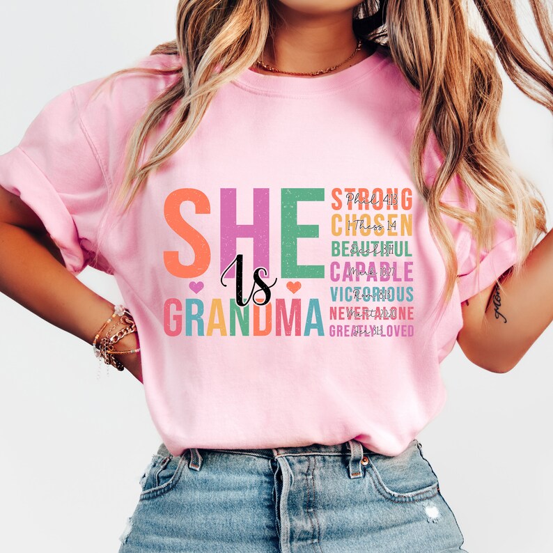 May include: Pink t-shirt with the text "She is Grandma" in rainbow colors. The text is stacked vertically with the words "Strong, Chosen, Beautiful, Capable, Victorious, Never Alone, Greatly Loved" in smaller font sizes.
