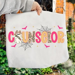 Pu&ograve; includere: Felpa bianca con la scritta "COUNSELOR" in lettere rosa e beige. La lettera "O" in "COUNSELOR" ha un motivo leopardato. La felpa ha un tema di Halloween con pipistrelli, ragnatele e un fantasma.