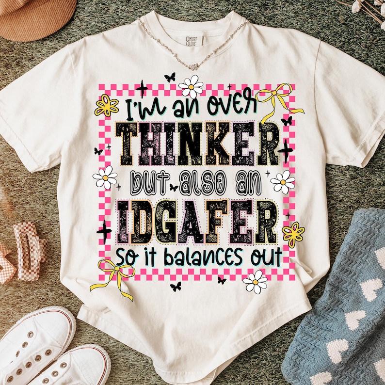 May include: Cream-colored t-shirt with the text "I'm an over thinker but also an IDGAF-er so it balances out." The design includes pink checkered borders, flowers, butterflies, and yellow bows. The shirt has a crew neck.