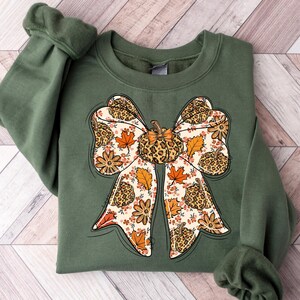 May include: An olive green sweatshirt featuring a large bow design. The bow is decorated with a pumpkin, leopard print, fall leaves, and flowers. The design is in shades of orange, brown, and cream.