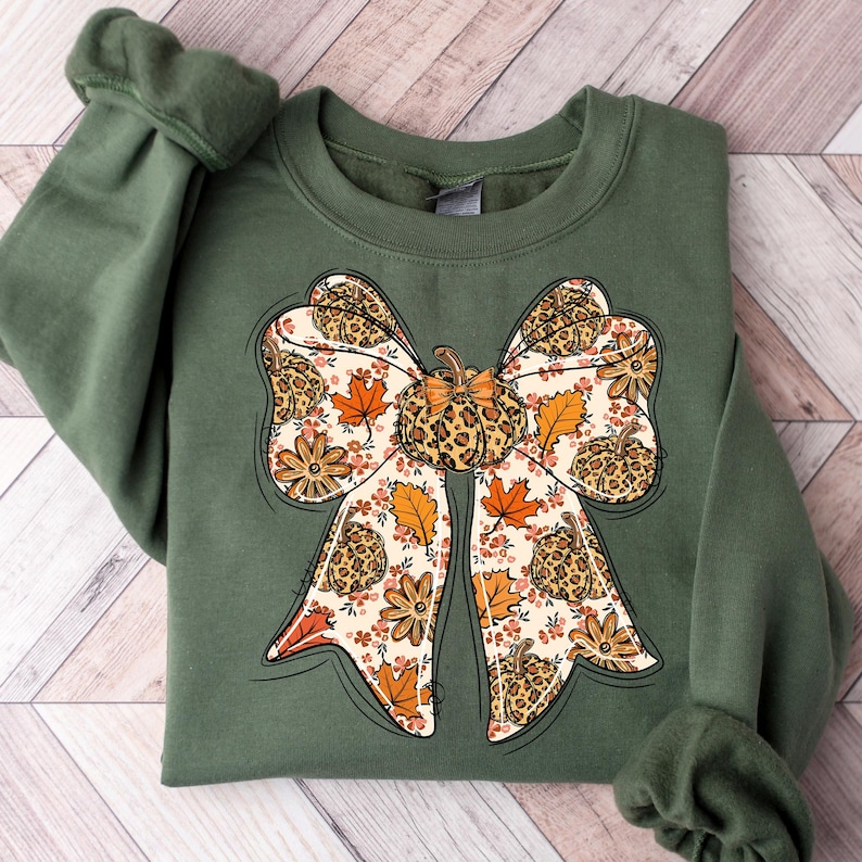 May include: An olive green sweatshirt featuring a large bow design. The bow is decorated with a pumpkin, leopard print, fall leaves, and flowers. The design is in shades of orange, brown, and cream.
