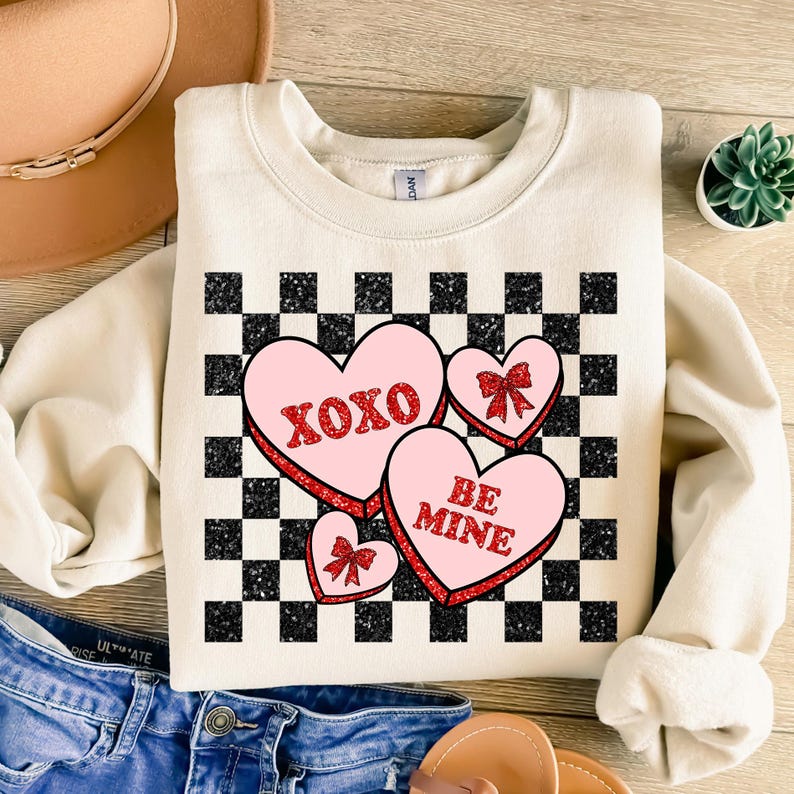 May include: A cream-colored sweatshirt with a black and white checkered pattern. The pattern features three pink hearts with red glitter accents. The hearts have the text "XOXO" and "BE MINE" on them.
