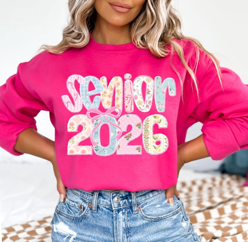 Puede incluir: Una sudadera rosa brillante con las palabras "Senior 2026" en un dise&ntilde;o floral y pastel. La sudadera es de cuello redondo y tiene mangas largas. El texto est&aacute; en una fuente decorativa.