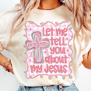 May include: Cream-colored t-shirt featuring a pink and white gingham design with a cross and the text "Let me tell you about my Jesus" in pink. The cross has a pink bow with blue and pink stripes.