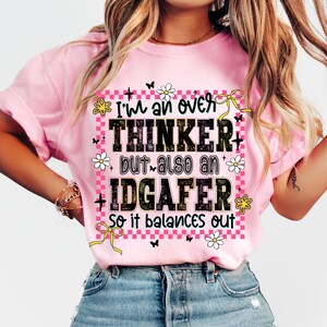 May include: Pink t-shirt with the text "I'm an over thinker but also an IDGAFer so it balances out." The text is in black with decorative flowers, bows, and butterflies. The shirt has short sleeves and a crew neck.