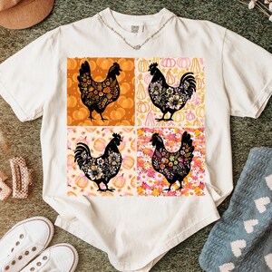 May include: White t-shirt featuring a four-panel design with black chicken silhouettes filled with floral patterns. The background panels include pumpkins and flowers in orange, pink, and cream colors. The shirt has a crew neck and short sleeves.