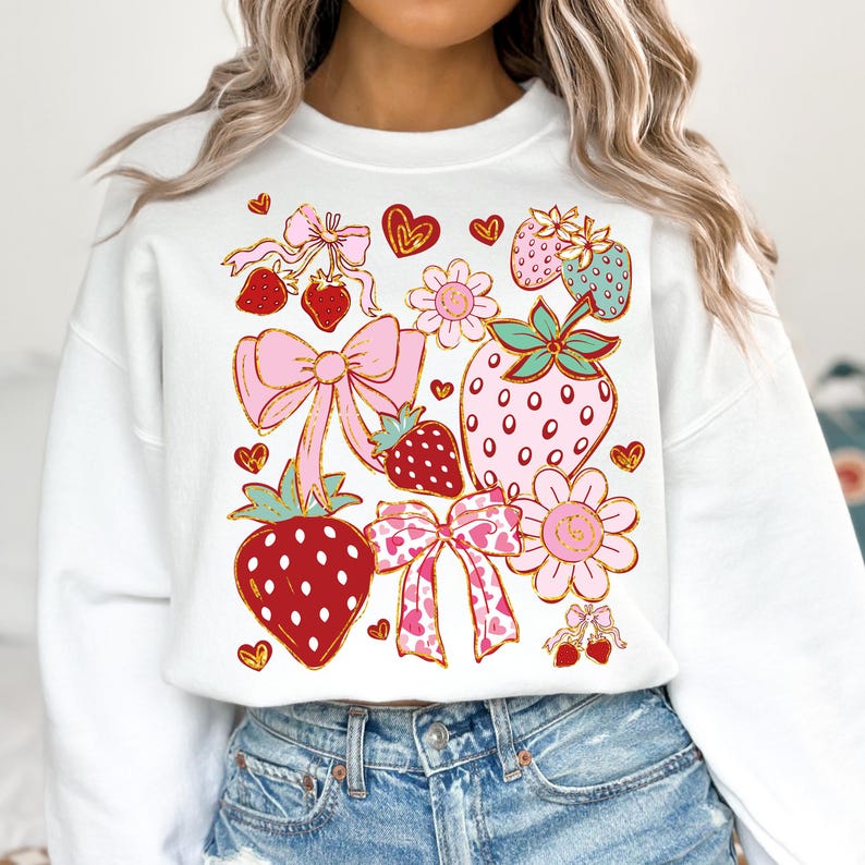 Op de afbeelding: Witte trui met een kleurrijke grafische print van aardbeien, strikken en bloemen. Het ontwerp bevat roze, rode, groene en gouden accenten.