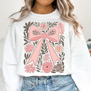 K&ouml;nnte beinhalten: Wei&szlig;es Sweatshirt mit einem gro&szlig;en rosa Schleifenmotiv, umgeben von rosa und grauen Blumen und gr&uuml;nen Bl&auml;ttern. Die Schleife ist der Mittelpunkt, mit detaillierten B&auml;ndern und einem Knoten. Das florale Design verleiht der Freizeitkleidung eine zarte Note.