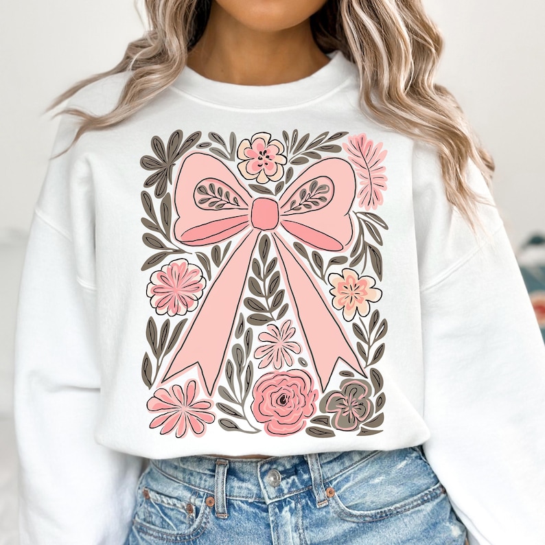 K&ouml;nnte beinhalten: Wei&szlig;es Sweatshirt mit einem gro&szlig;en rosa Schleifenmotiv, umgeben von rosa und grauen Blumen und gr&uuml;nen Bl&auml;ttern. Die Schleife ist der Mittelpunkt, mit detaillierten B&auml;ndern und einem Knoten. Das florale Design verleiht der Freizeitkleidung eine zarte Note.