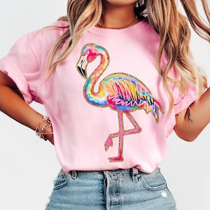 May include: Pink sweatshirt featuring a colorful flamingo design. The flamingo has a rainbow-colored body with gold glitter accents. The bird is standing on one leg. The sweatshirt is a solid pink color.